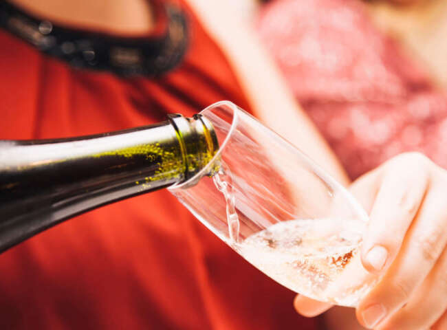 The hands of an unknown woman wearing a red dress holding a glass as she fills it with cava from a green glass bottle.
