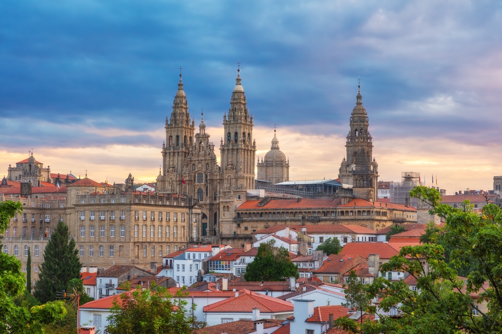 Aerial view of Santiago de Compostela at sunrise in Galicia, Spain, featuring the grand Gothic Cathedral rising above historic stone buildings, with warm golden light illuminating the city’s rooftops and narrow streets.