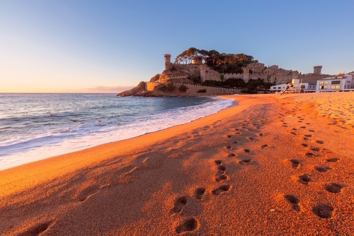 Sunrise on the beach with waves washing the shore and a historical fortress in Tossa de Mar, Spain.