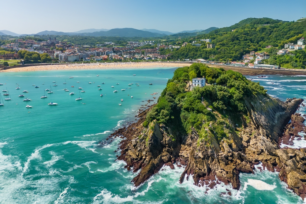 Aerial view of San Sebastián Bay in Donostia, Basque Country, Spain, featuring vibrant turquoise waters, the city’s scenic coastline, and Santa Clara Island with its historic lighthouse, captured from above on a sunny summer day.